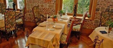 Un comedor con paredes de ladrillo, ventanas amplias y suelo de madera. Las mesas están preparadas con platos, vasos y cubiertos.