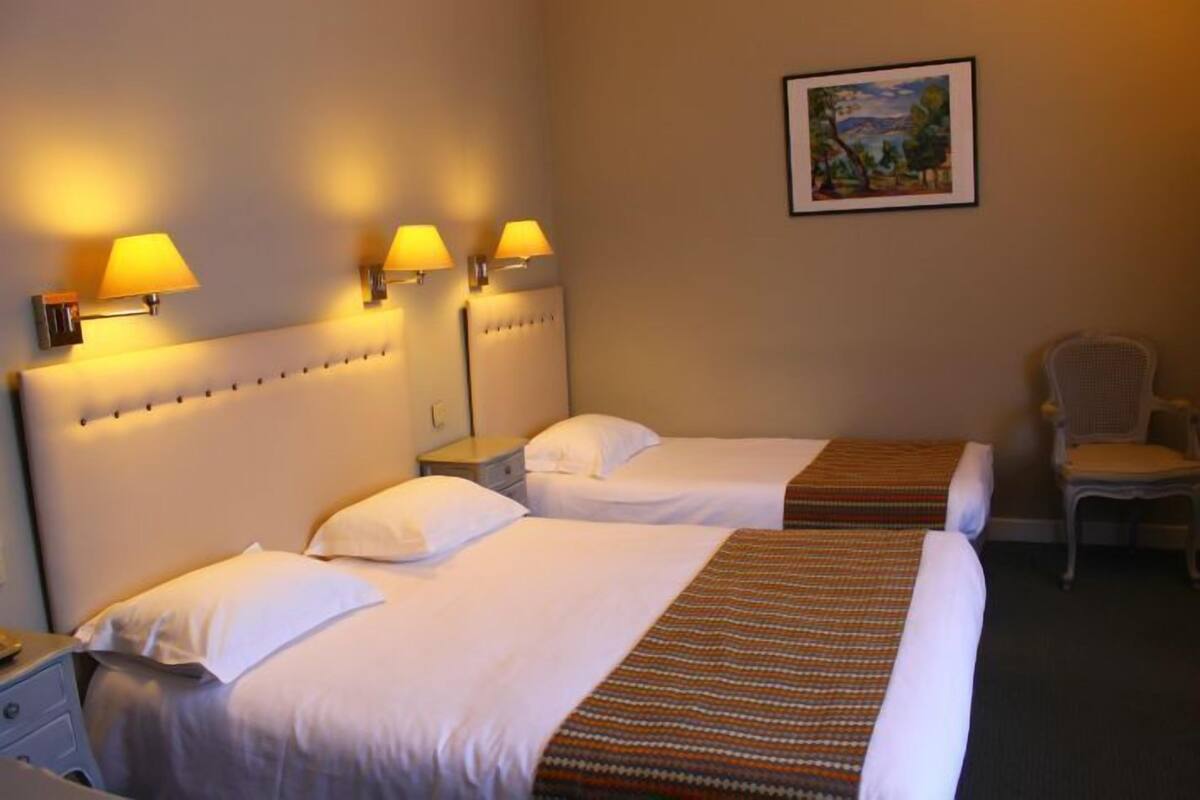 Ein Hotelzimmer mit zwei Betten, jeweils mit weißer Bettwäsche und einem gemusterten Überwurf. An beiden Seiten der Betten befinden sich Wandlampen, und an der Wand hängt ein gerahmtes Gemälde.