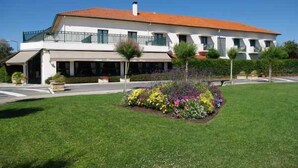 A two-story building with a red-tiled roof, a balcony on the upper floor, and a well-maintained garden with various flowers. - Hotel des Pins (Soulac-sur-Mer)