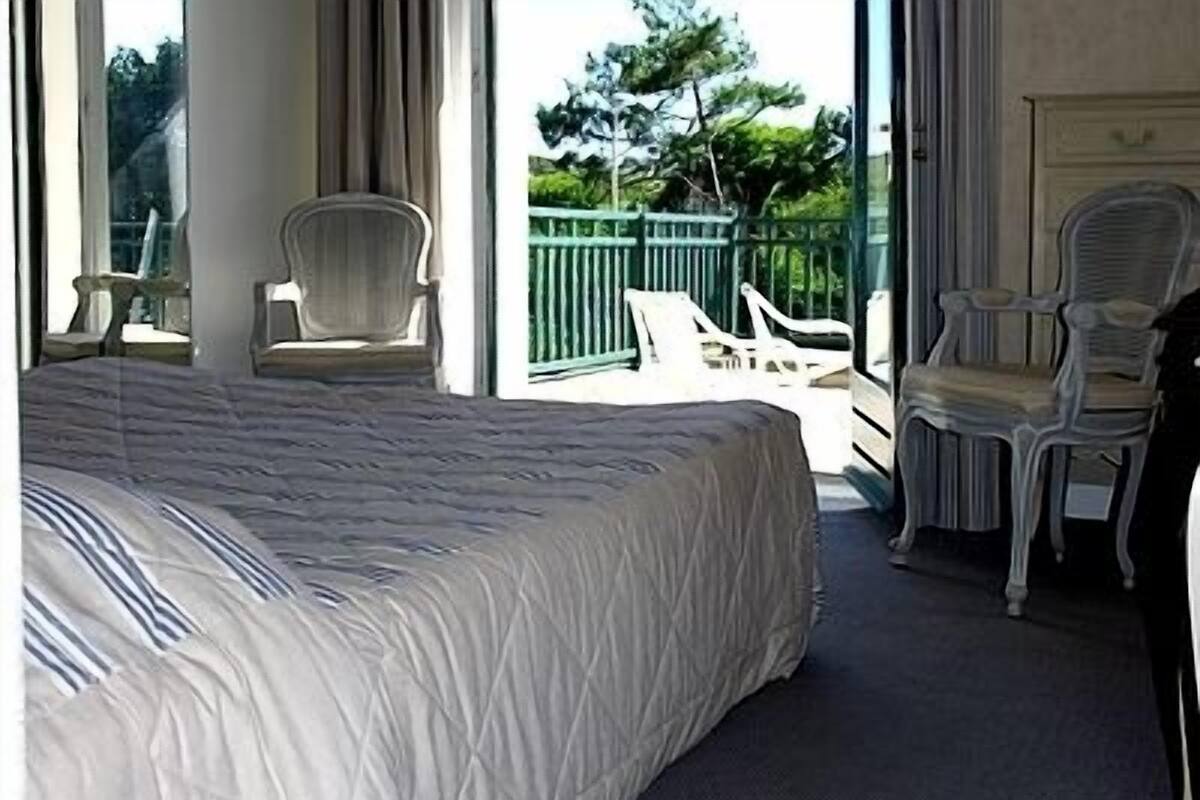 Ein Hotelzimmer mit einem großen Bett, zwei Stühlen, einem Balkon mit grüner Geländer und Blick auf Bäume und einen Zaun.