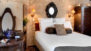 A bedroom with a stone wall, a bed with white linens, a wooden dresser, and a mirror with a decorative frame. - Hôtel Saint Martin - La Maison Younan (Saint-Maixent-l'Ecole)