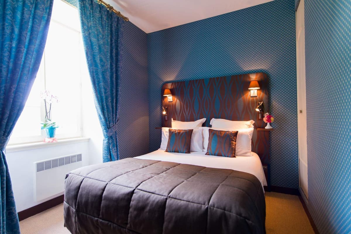 Une chambre d’hôtel avec un grand lit, une tête de lit dotée de lampes murales, une fenêtre avec des rideaux et un radiateur.