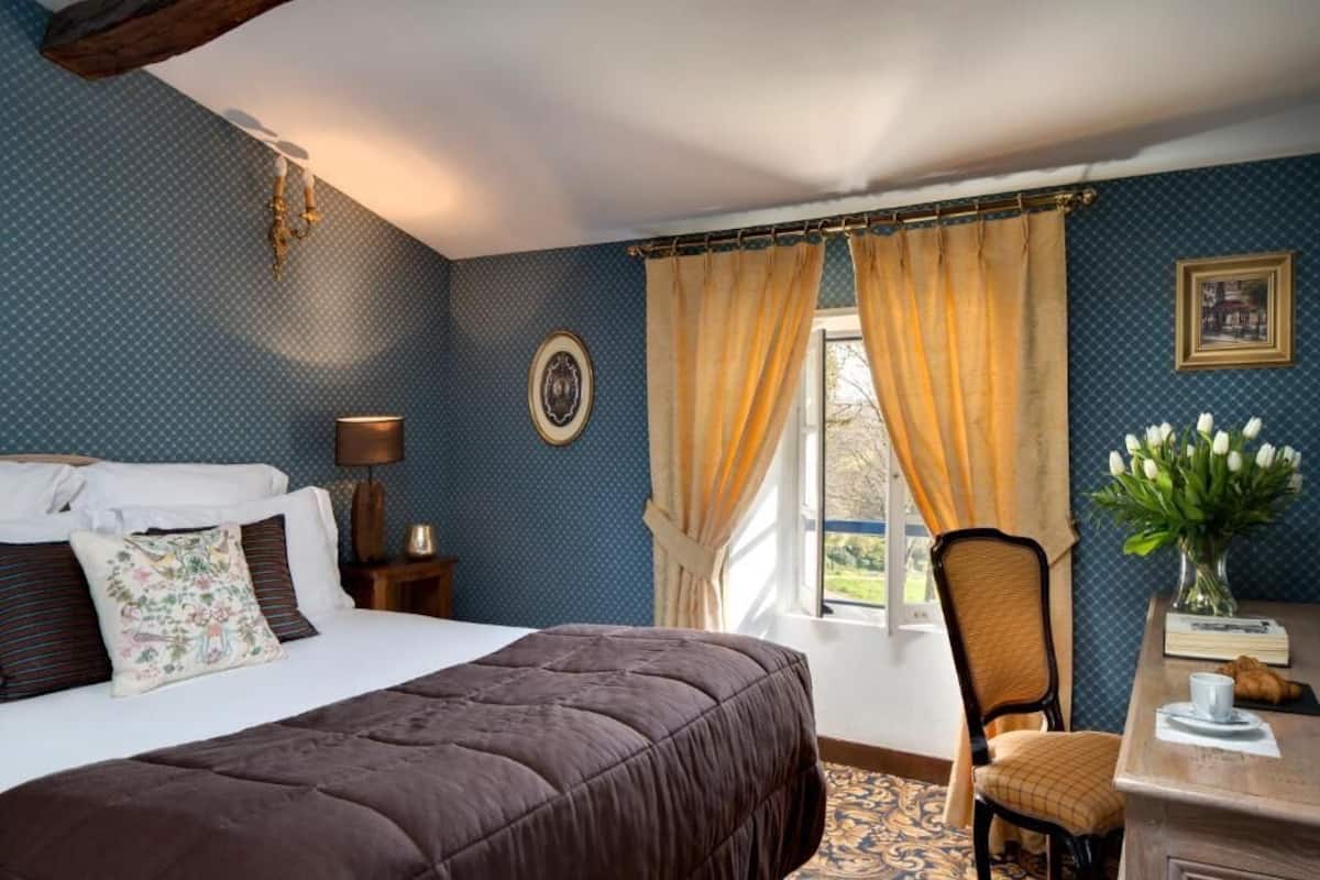 Une chambre avec un grand lit, un bureau avec une chaise, une fenêtre avec des rideaux et un vase rempli de fleurs.