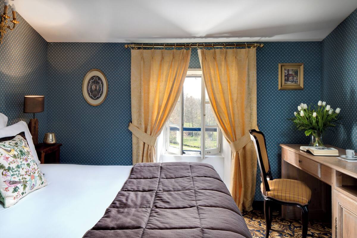 Une chambre à coucher avec un lit, un bureau, une chaise, une fenêtre avec des rideaux, une lampe et un vase avec des fleurs.