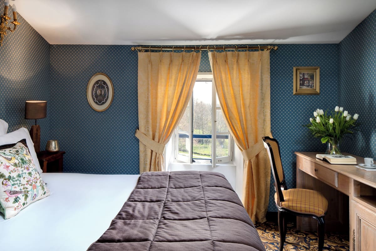 A bedroom with a bed, a desk, a chair, a window with curtains, a lamp, and a vase with flowers.
