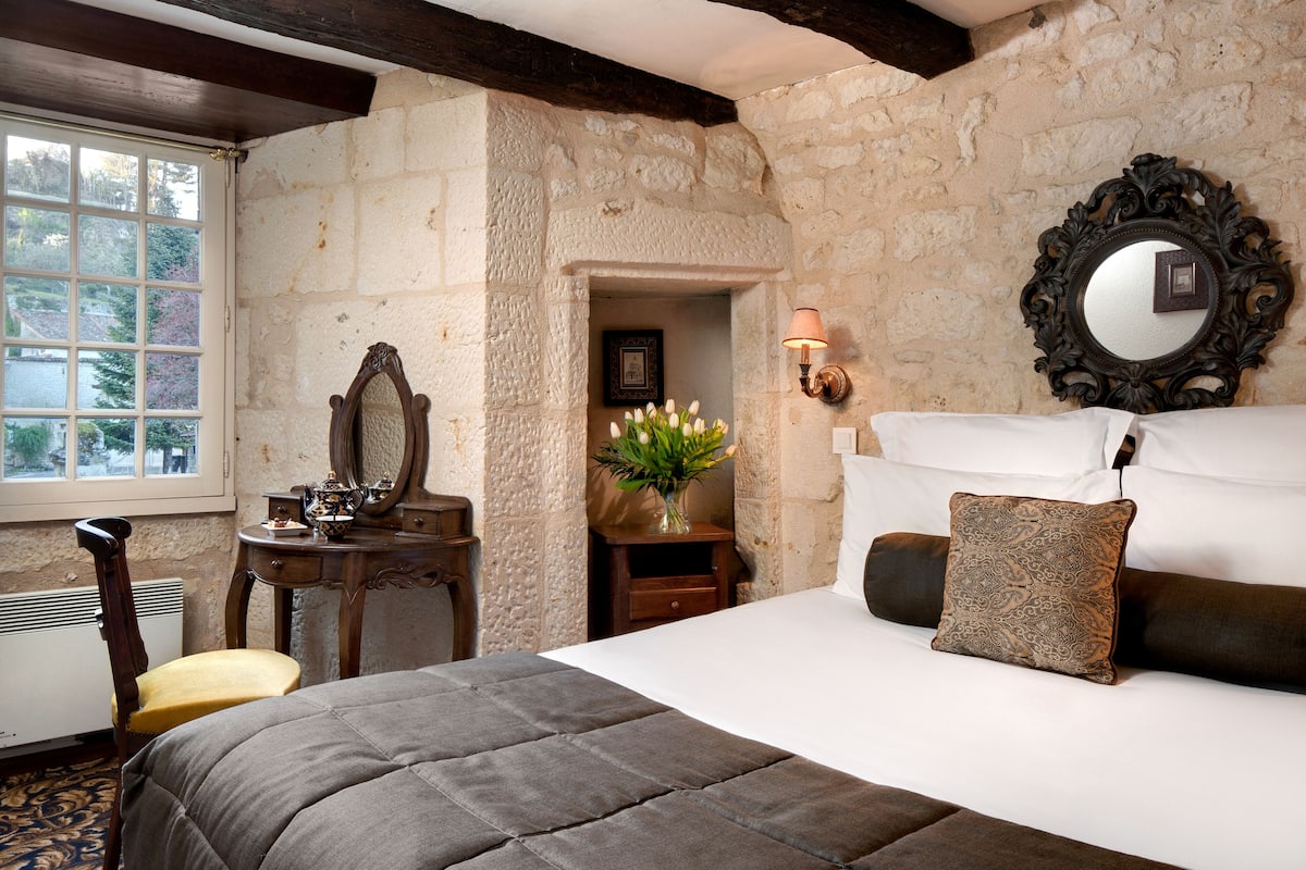 Une chambre avec un mur en pierre, un lit recouvert d’une courtepointe, une commode en bois, un miroir et une fenêtre donnant sur un paysage agréable.