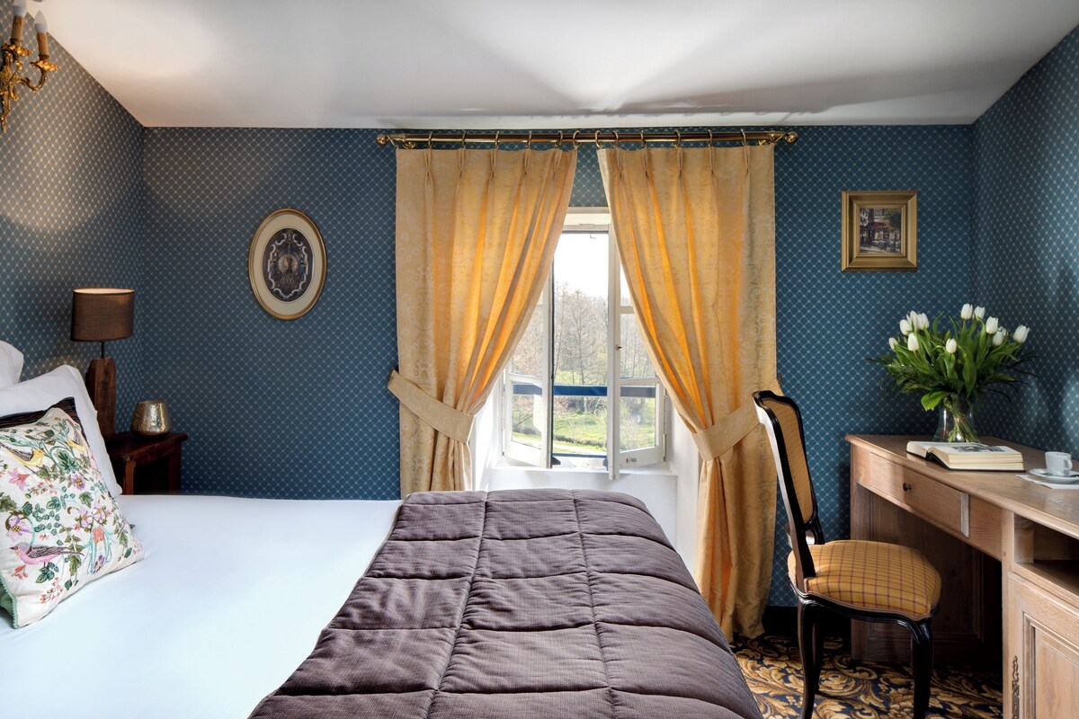 A bedroom with a bed, a desk, a chair, a window with curtains, a lamp, and a vase with flowers.