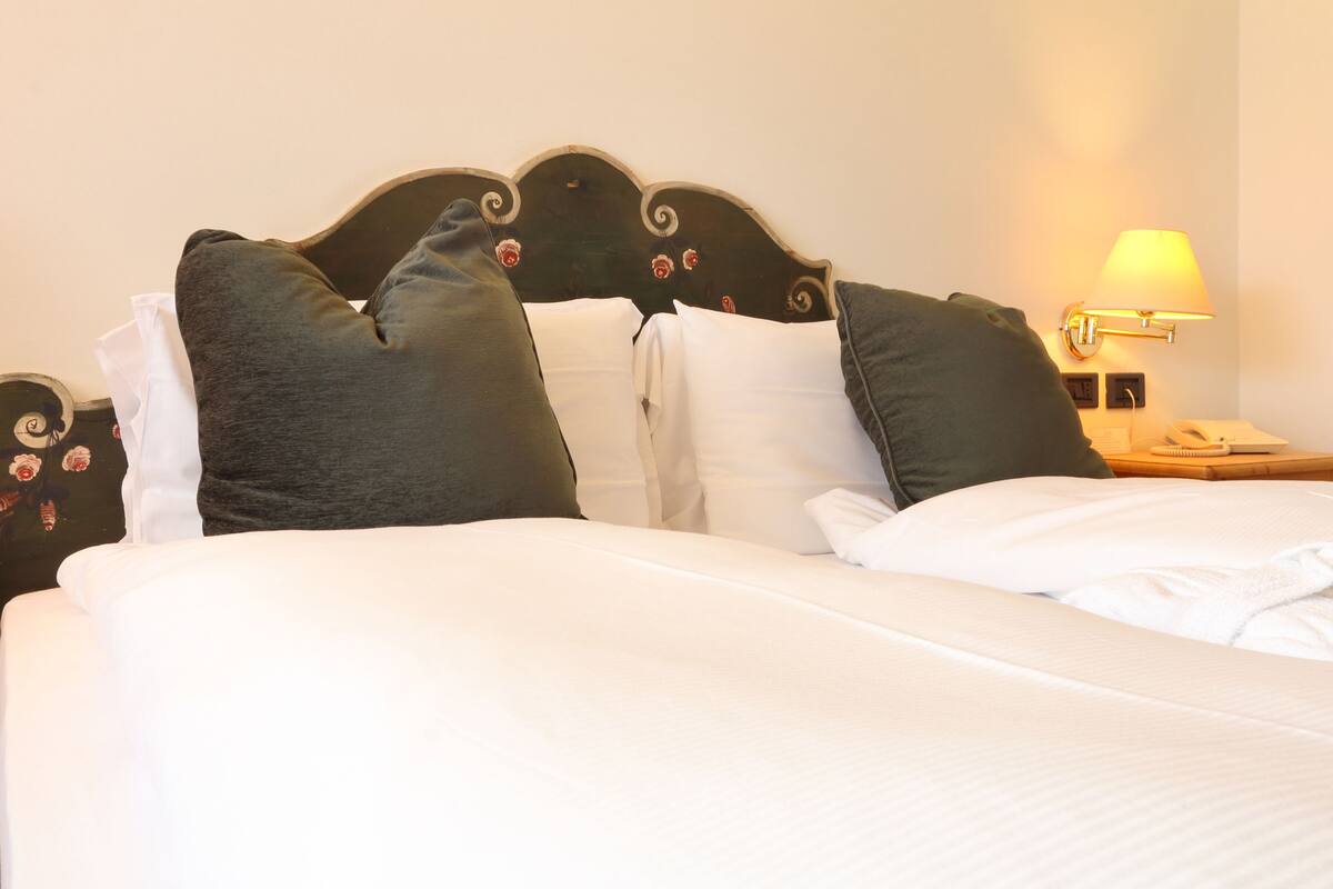 Una cama bien hecha con sábanas blancas y almohadas oscuras, un cabecero de madera con detalles florales, y una lámpara de noche sobre una mesita.