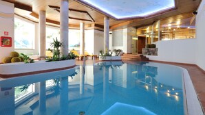 An indoor swimming pool with clear blue water, surrounded by wooden pillars, potted plants, and a view of the outdoors.