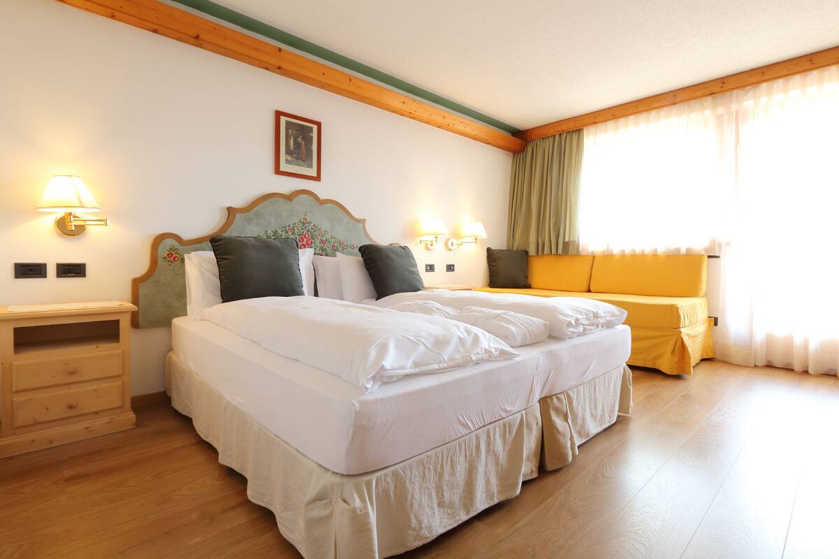 Una cama doble con sábanas blancas, dos mesitas de noche con lámparas, un tocador de madera y un sofá amarillo.