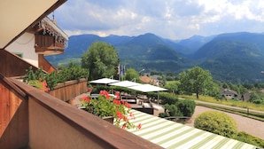 A balcony with a wooden railing overlooking a mountainous landscape. There are outdoor seating areas with umbrellas and potted plants.