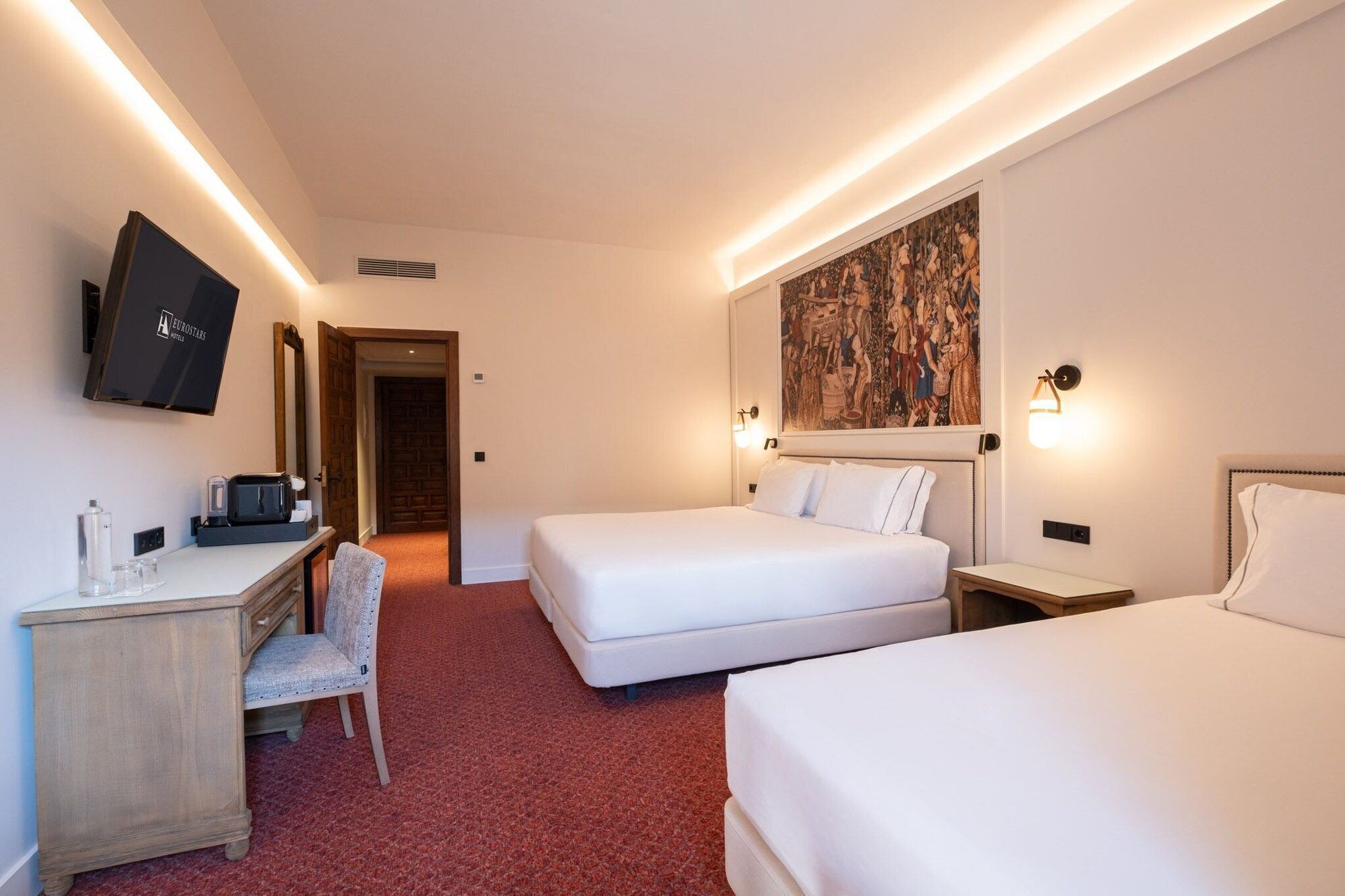 Habitación de hotel con escritorio, dos camas, televisor, un cuadro y una alfombra roja.