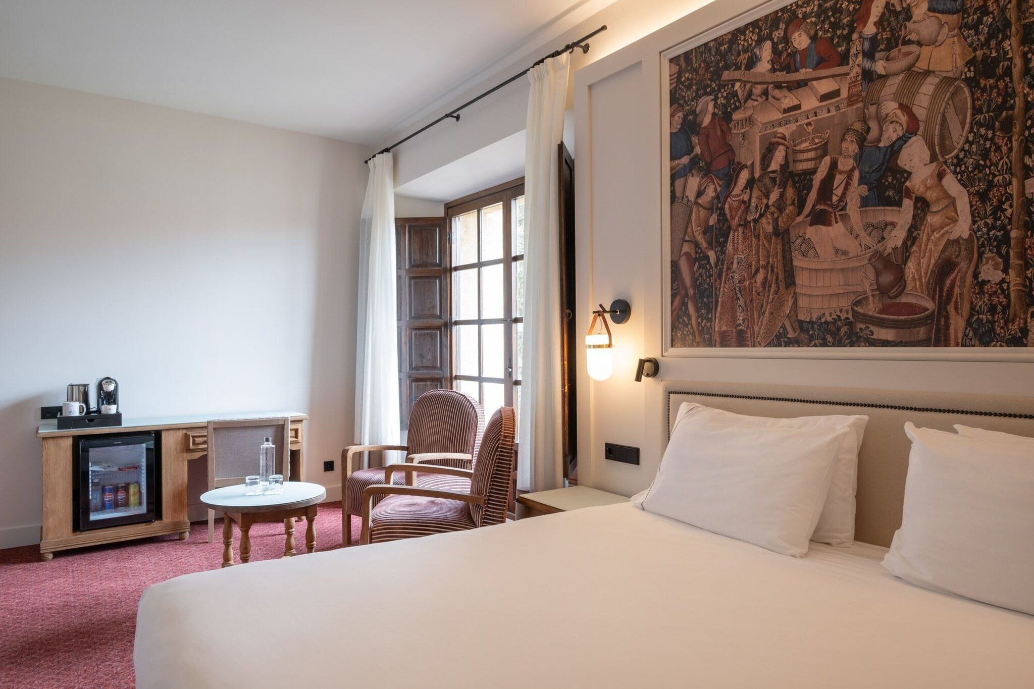 Habitación de hotel con cama, escritorio con refrigerador, una silla, una mesita y un tapiz grande en la pared.