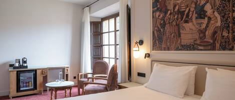 A hotel room with a bed, a desk with a refrigerator, a chair, a table, and a large tapestry on the wall.