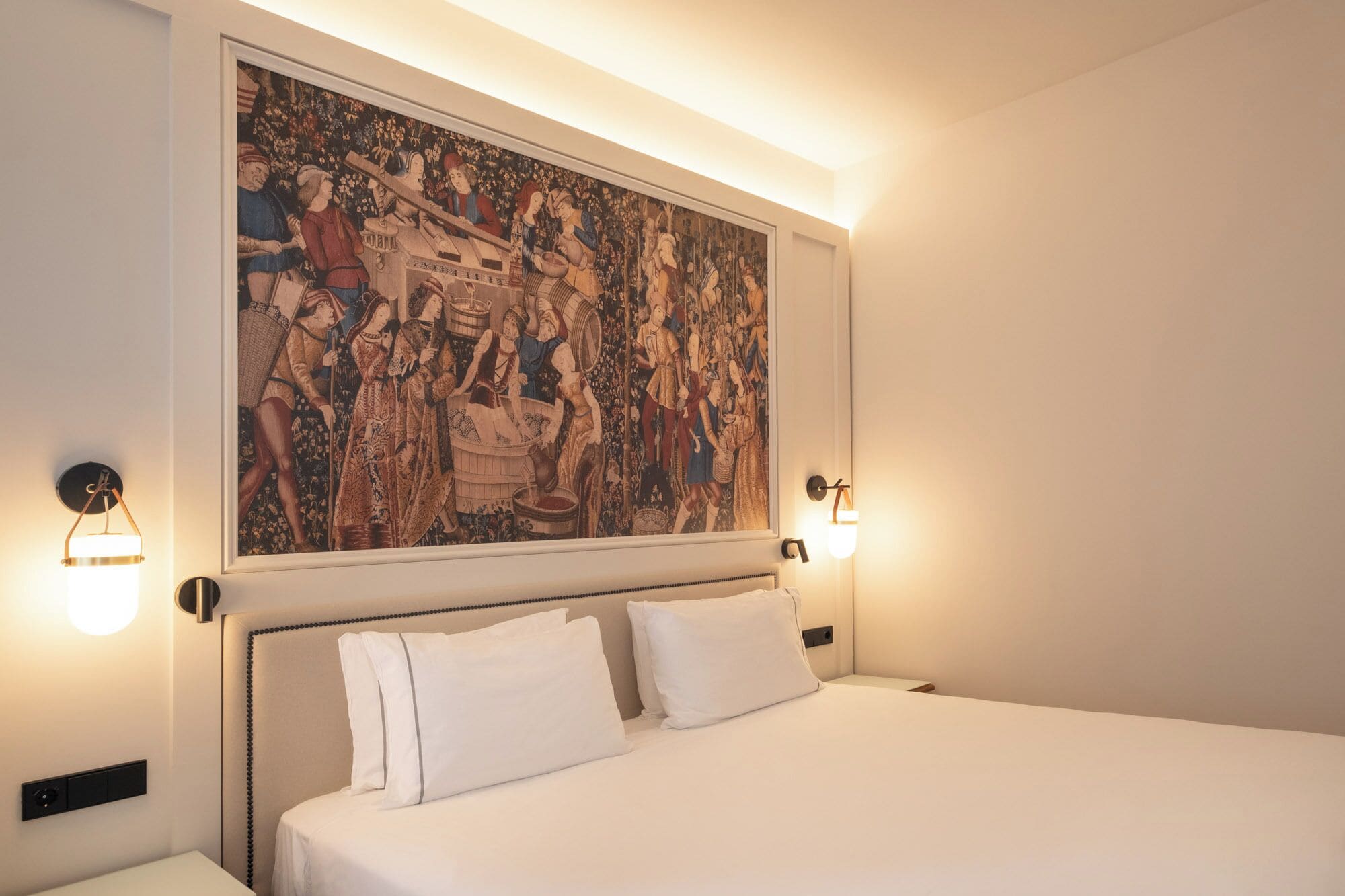 Een hotelkamer met een groot wandtapijt, een bed met witte lakens en wandlampen.