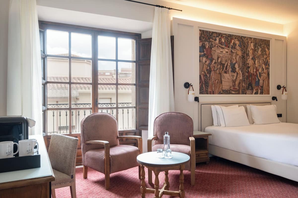 Ein Hotelzimmer mit einem Bett, zwei gestreifte Sessel, einem kleinen runden Tisch mit einer Wasserflasche, einer Kaffeemaschine und einem großen Fenster mit Blick auf Gebäude.