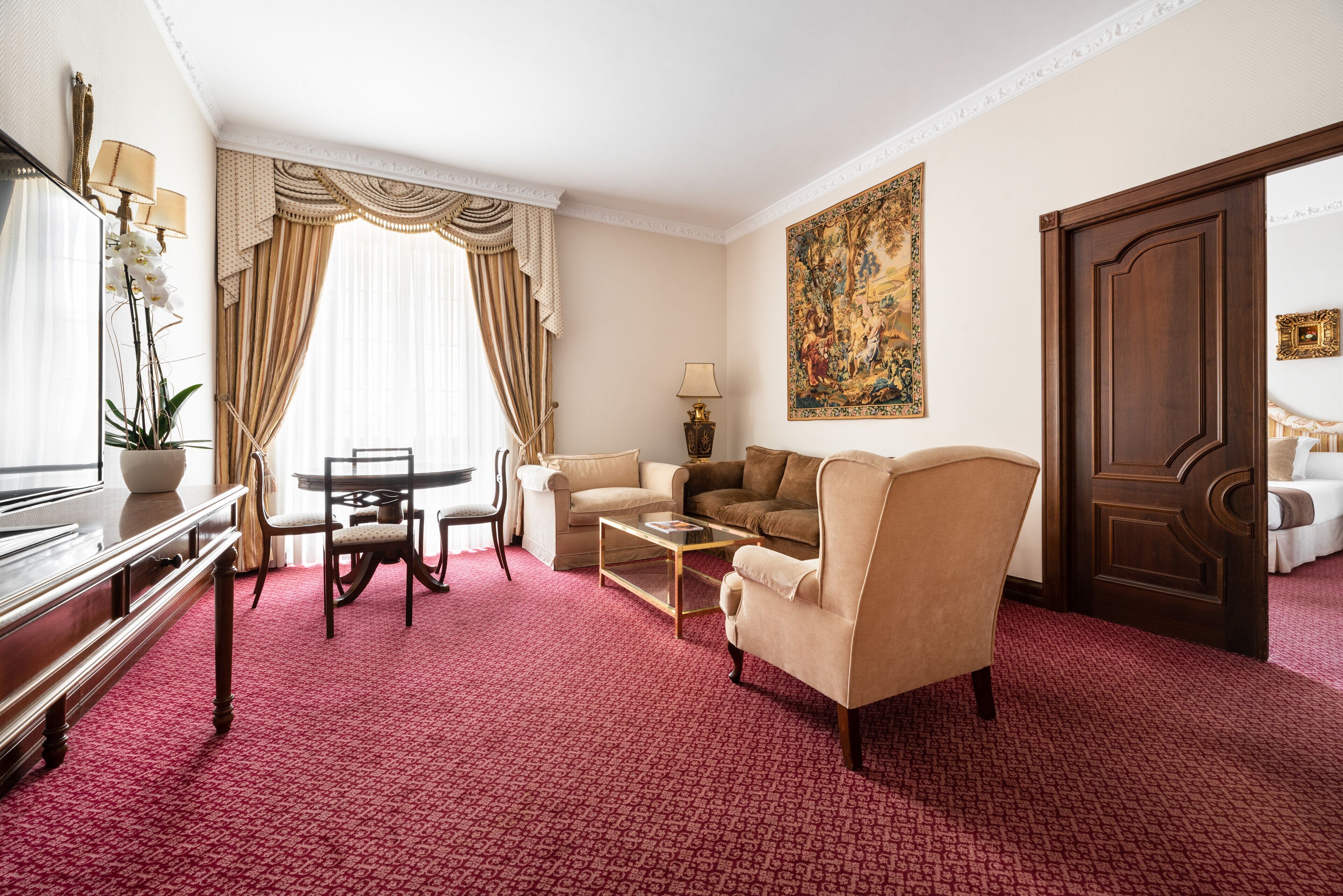 A hotel room with a red carpet, a sofa, a chair, a coffee table, a desk with a lamp, and a large painting on the wall.