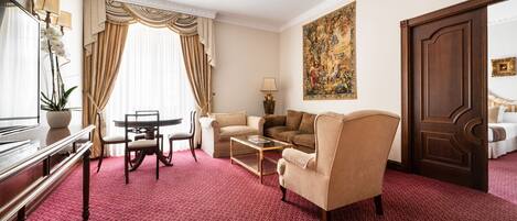 A hotel room with a red carpet, a sofa, a chair, a coffee table, a desk with a lamp, and a large painting on the wall.