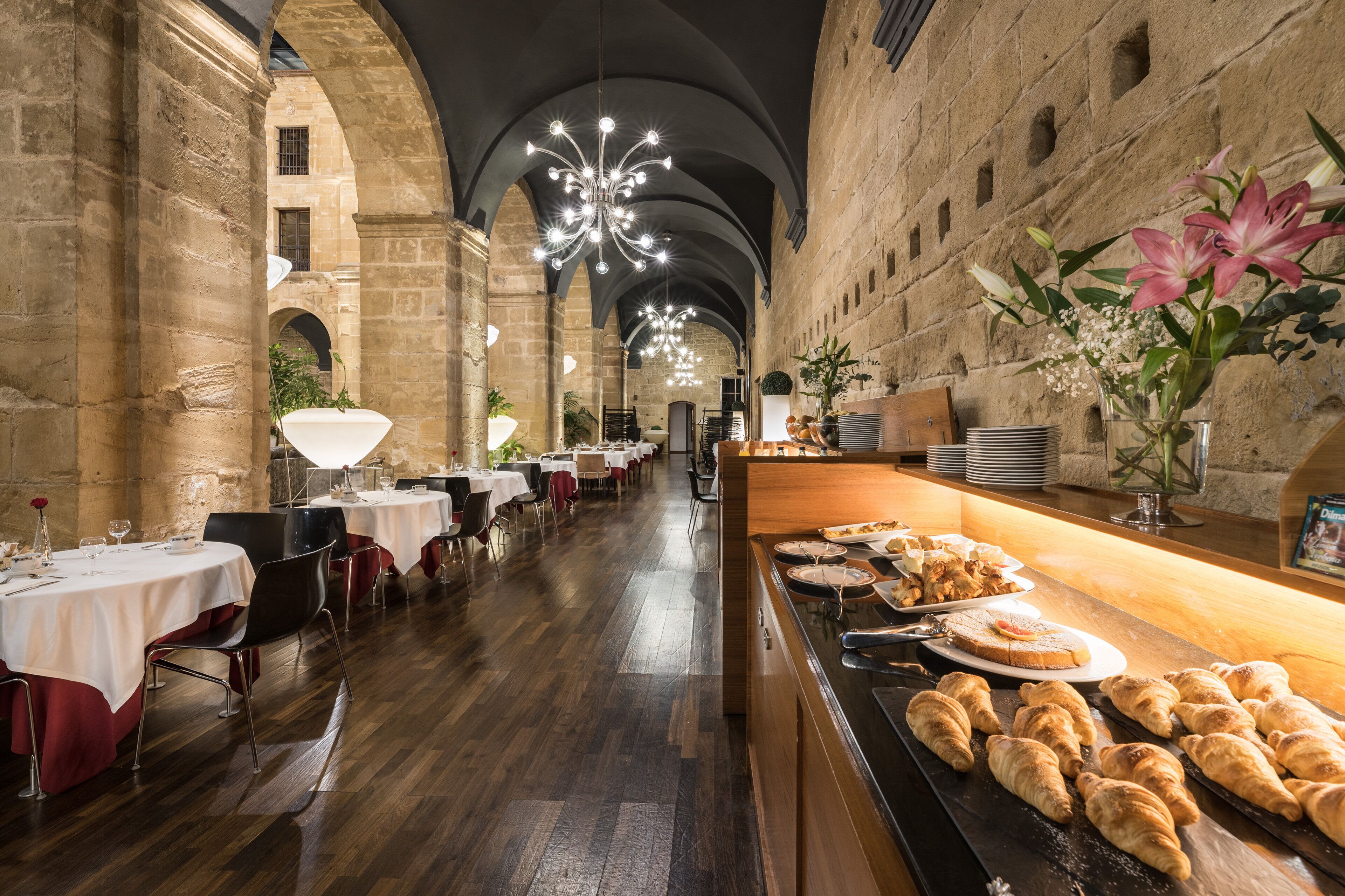 A spacious dining area with high ceilings and stone walls, featuring tables set for dining and a buffet with pastries.