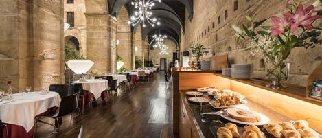 A spacious dining area with high ceilings and stone walls, featuring tables set for dining and a buffet with pastries.