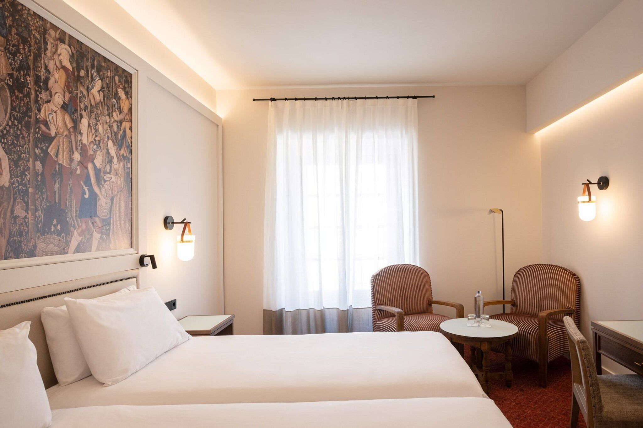 A hotel room with a large bed, two chairs, a small table, and a painting on the wall.