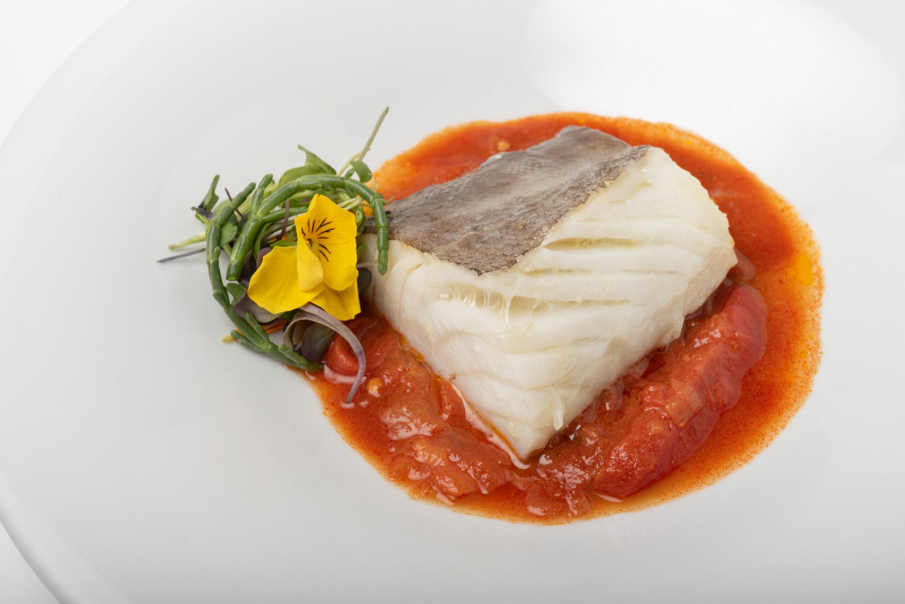 Un plato blanco con un trozo de pescado, salsa roja y una flor amarilla.