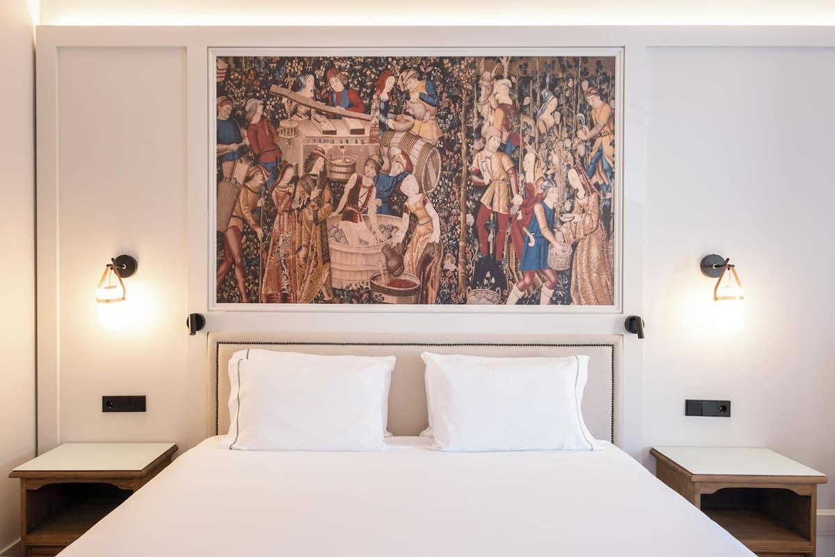 Ein Schlafzimmer mit einem großen Wandteppich im mittelalterlichen Stil, zwei Nachttischen und einem Bett mit weißen Leinen.
