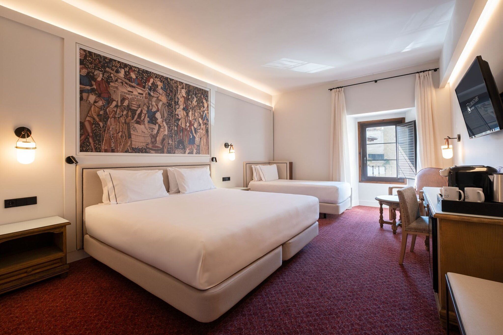 A hotel room with a large bed, a smaller bed, a desk with a chair, a television, and a painting on the wall.