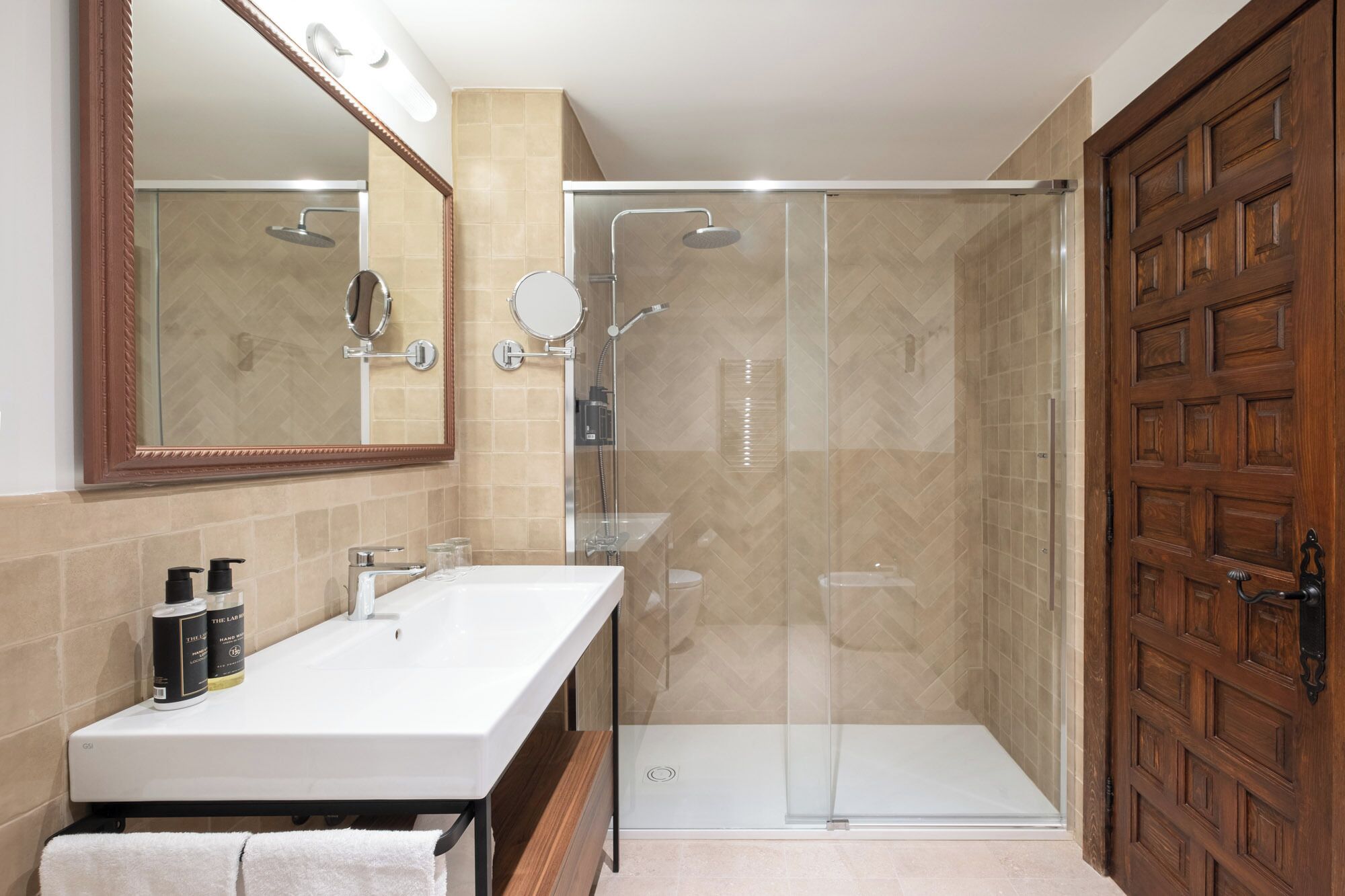A bathroom with a double sink, a mirror, a shower enclosure, and a wooden door.