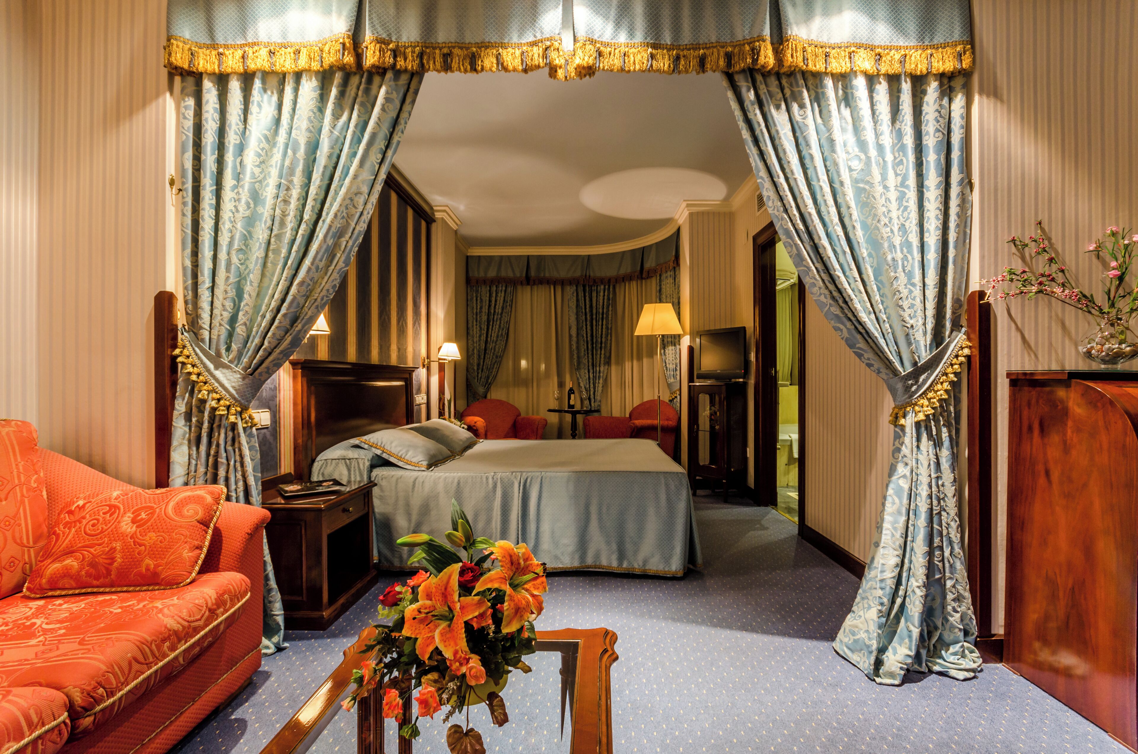 A hotel room with a bed, a sofa, a nightstand, a lamp, and a vase with flowers.