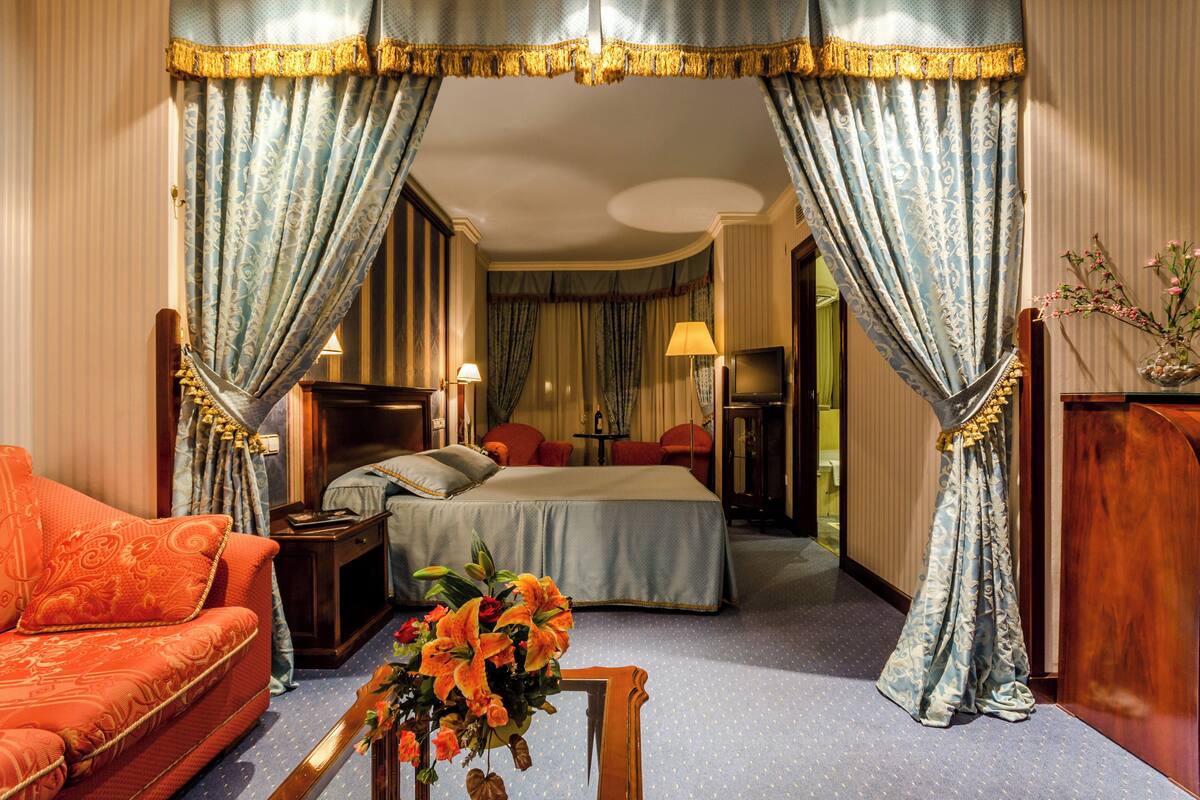 A hotel room with a bed, a sofa, a nightstand, a lamp, and a vase with flowers.