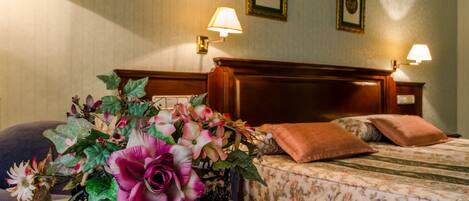 Una habitación de hotel con cama, mesita de noche, lámpara, un cuadro en la pared y un arreglo floral sobre la mesita.