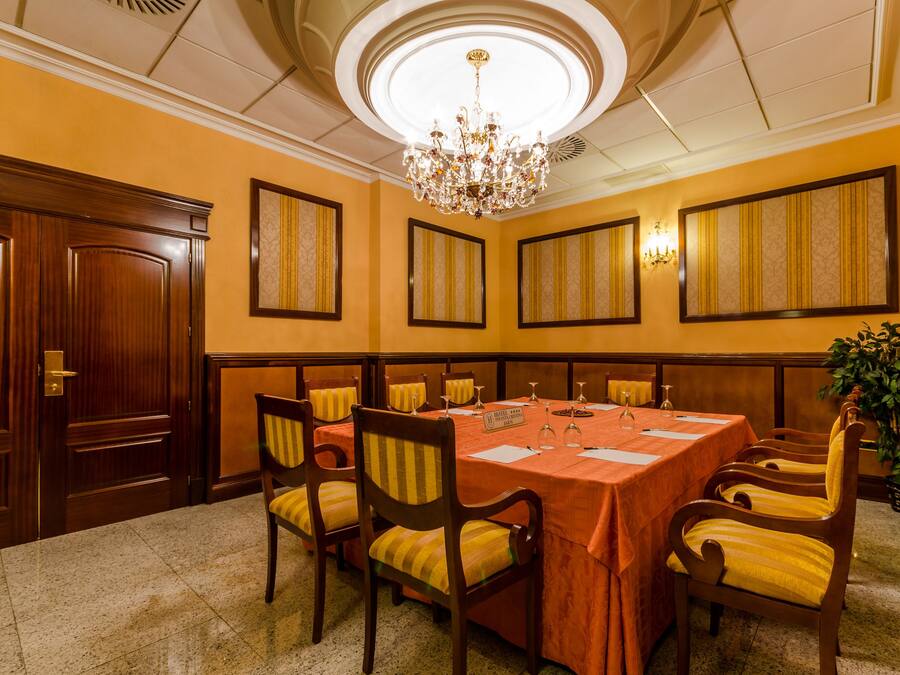 A conference room with a large oval table, wooden chairs with yellow cushions, a chandelier, and framed panels on the walls.