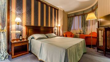 A hotel room with a large bed, two bedside tables with lamps, an orange armchair, a floor lamp, and a television on a cabinet.