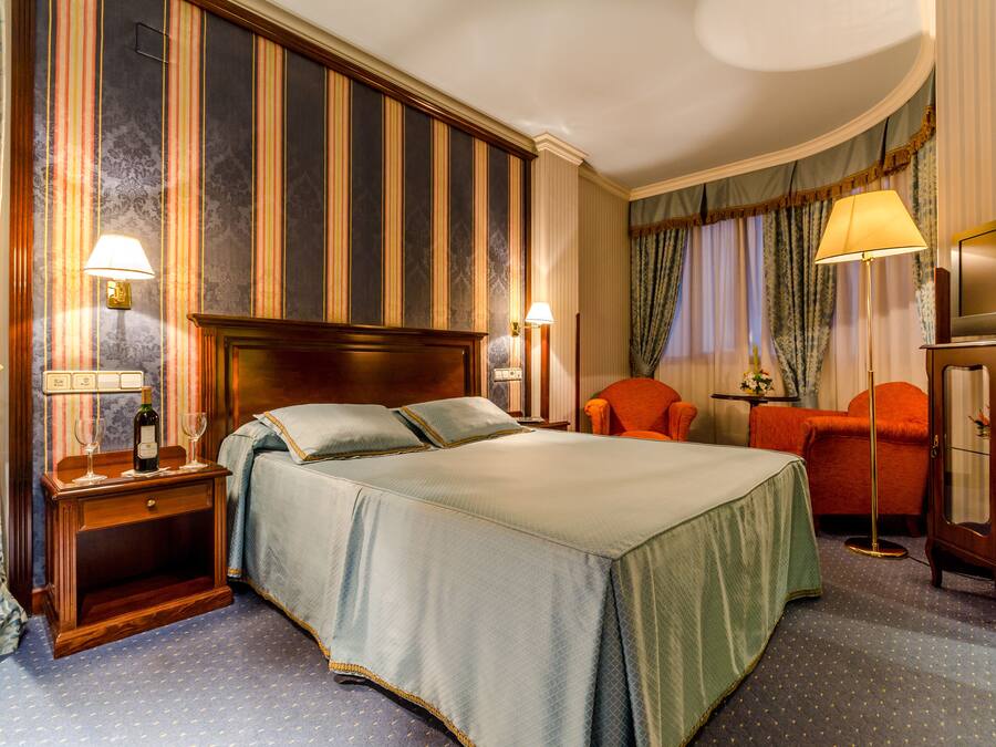 A hotel room with a large bed, two bedside tables with lamps, an orange armchair, a floor lamp, and a television on a cabinet.