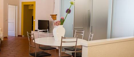 A dining area with a round table, six chairs, a white vase with flowers, and a television on a stand.