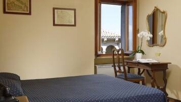 Habitación de hotel con cama, escritorio con silla, ventana con vistas a edificios y espejo en la pared.