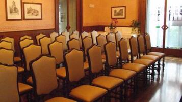 Un salón de conferencias con filas de sillas, cuadros en las paredes y un ventanal con persianas.