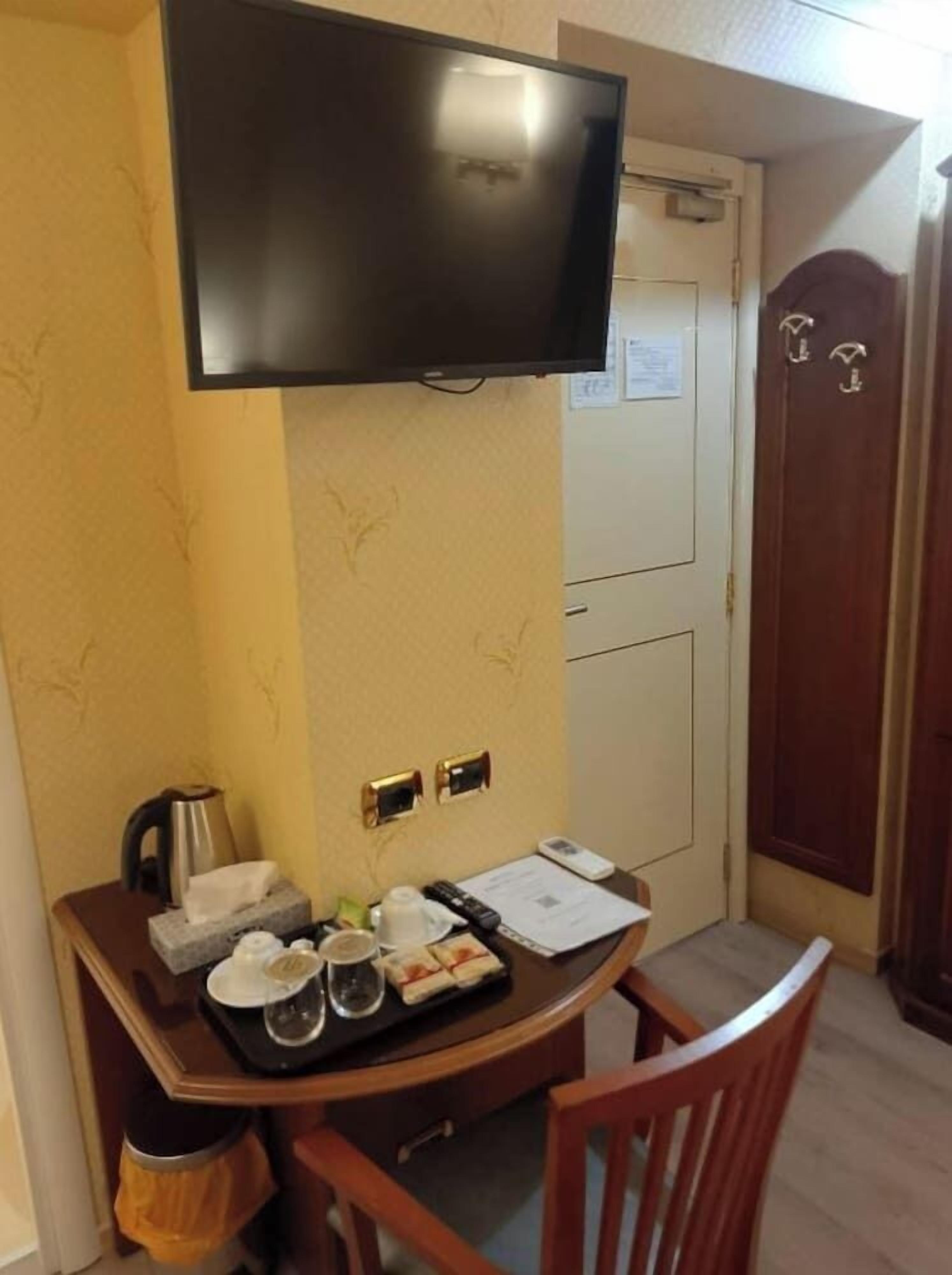A hotel room with a TV mounted on the wall, a desk with a kettle, cups, and a tray of food, and a wardrobe with a mirror.