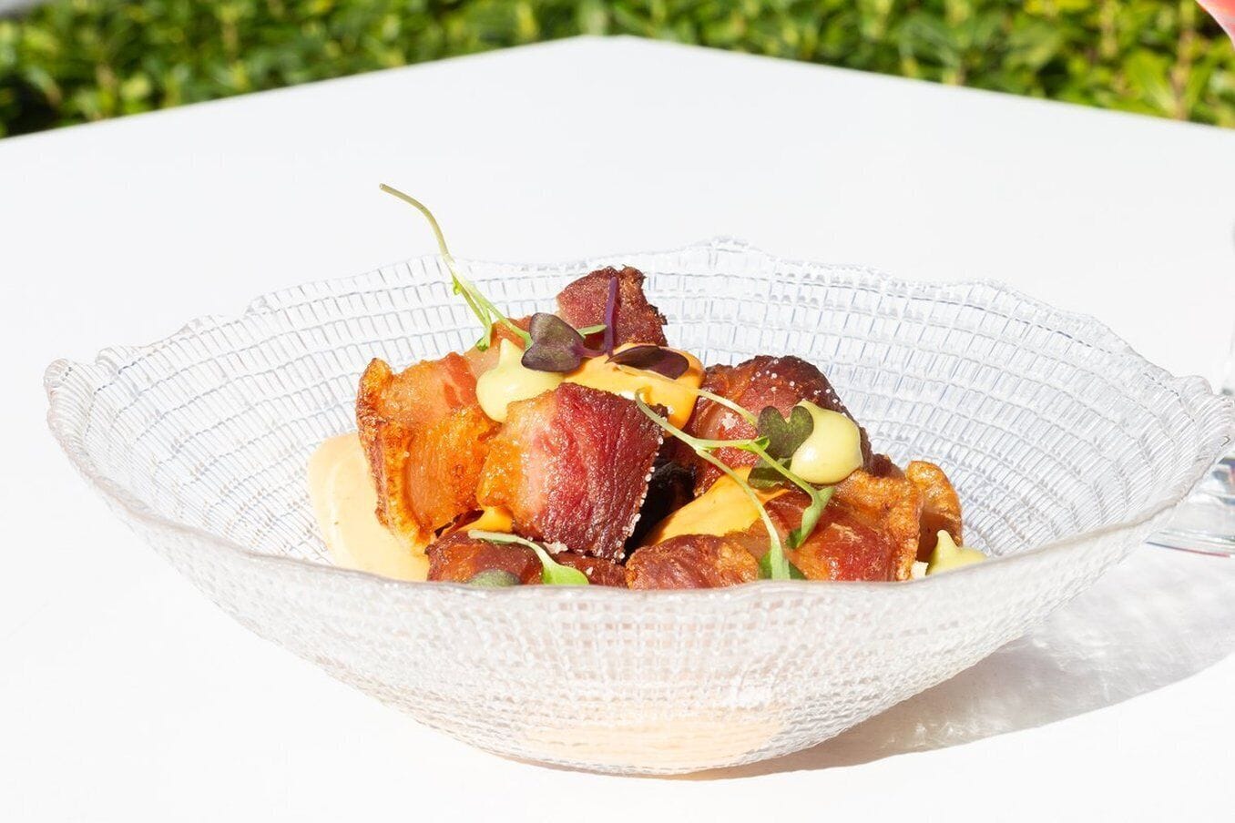 Un tazón de vidrio transparente con un plato de tocino, vegetales de hoja verde y salsa cremosa, sobre una mesa blanca, con un jardín verde de fondo.