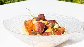 Un tazón de vidrio transparente con un plato de tocino, vegetales de hoja verde y salsa cremosa, sobre una mesa blanca, con un jardín verde de fondo.