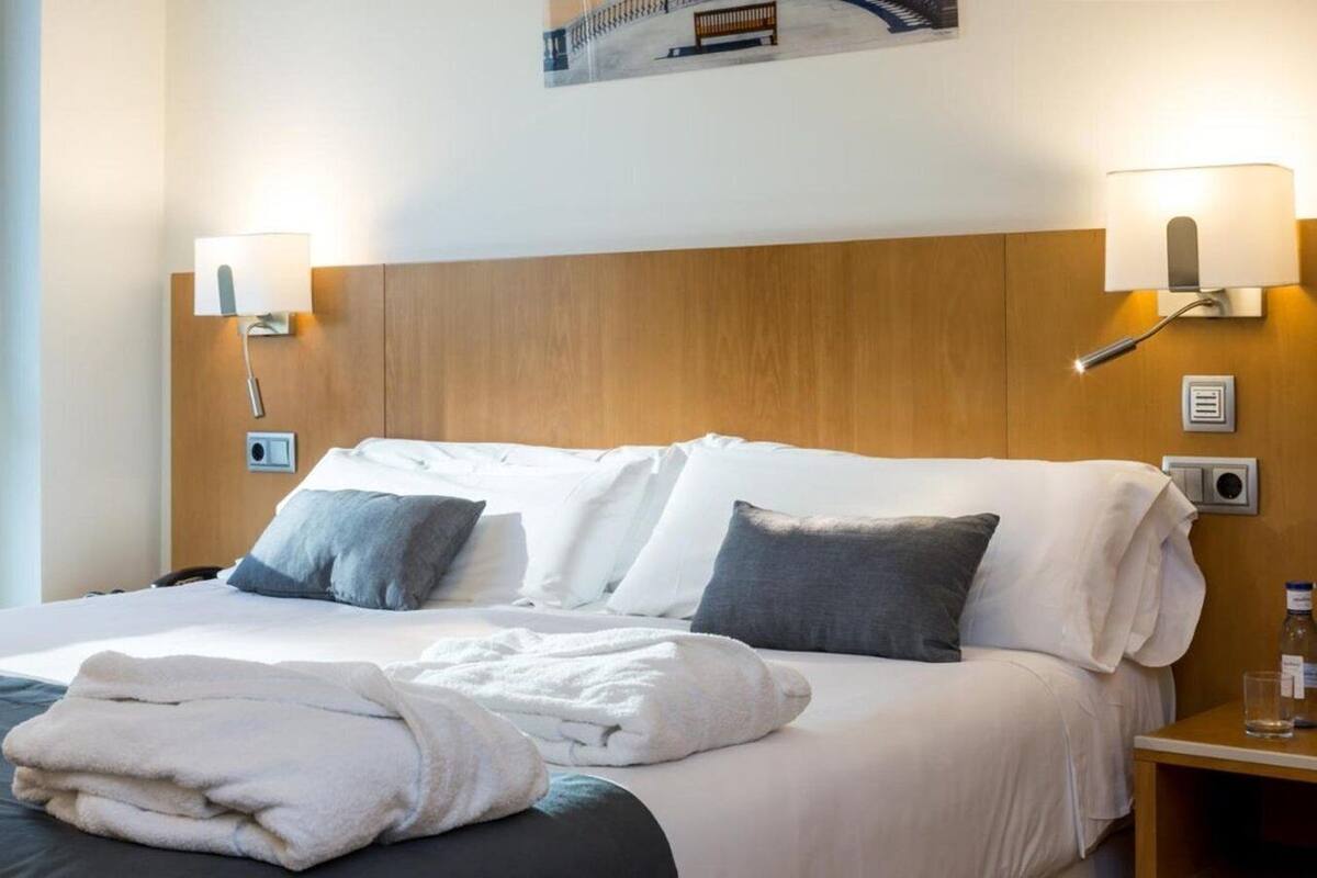 A hotel room with a wooden headboard, two bedside lamps, a bed with white linens, two pillows, a bedside table with a glass and a bottle, and a wall-mounted thermostat.