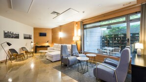 A modern hotel lobby with a sofa, armchairs, a coffee table, a bookshelf, and a large window offering a view of greenery.