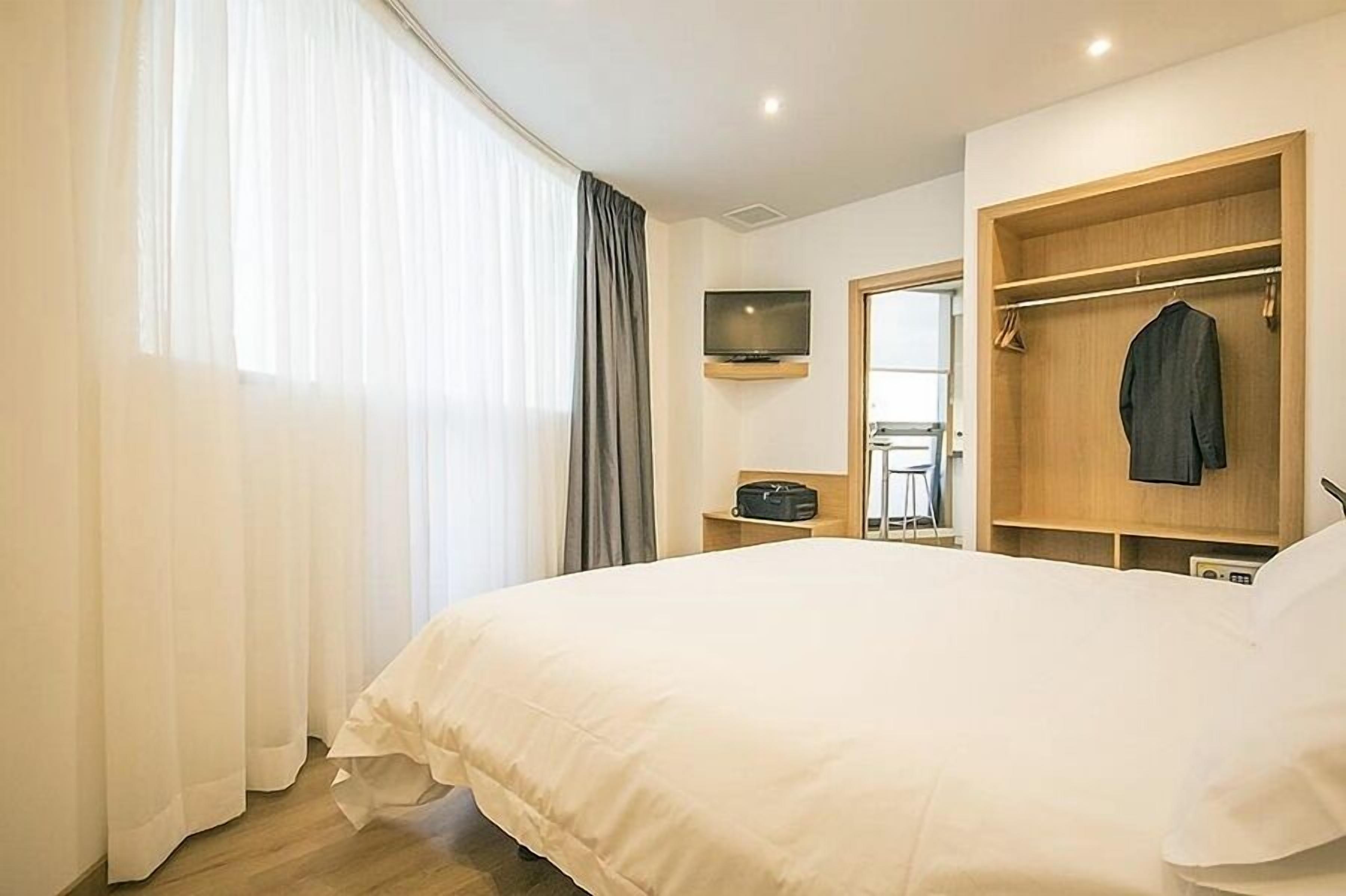 A hotel room with a large bed, a TV mounted on the wall, a wooden wardrobe, and a small table with a radio.