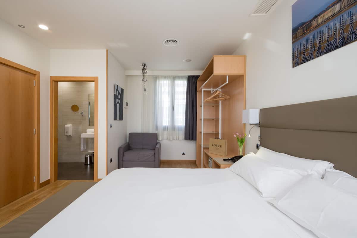 A hotel room with a bed, a desk with a lamp, a chair, a wardrobe, and a bathroom visible through an open door.