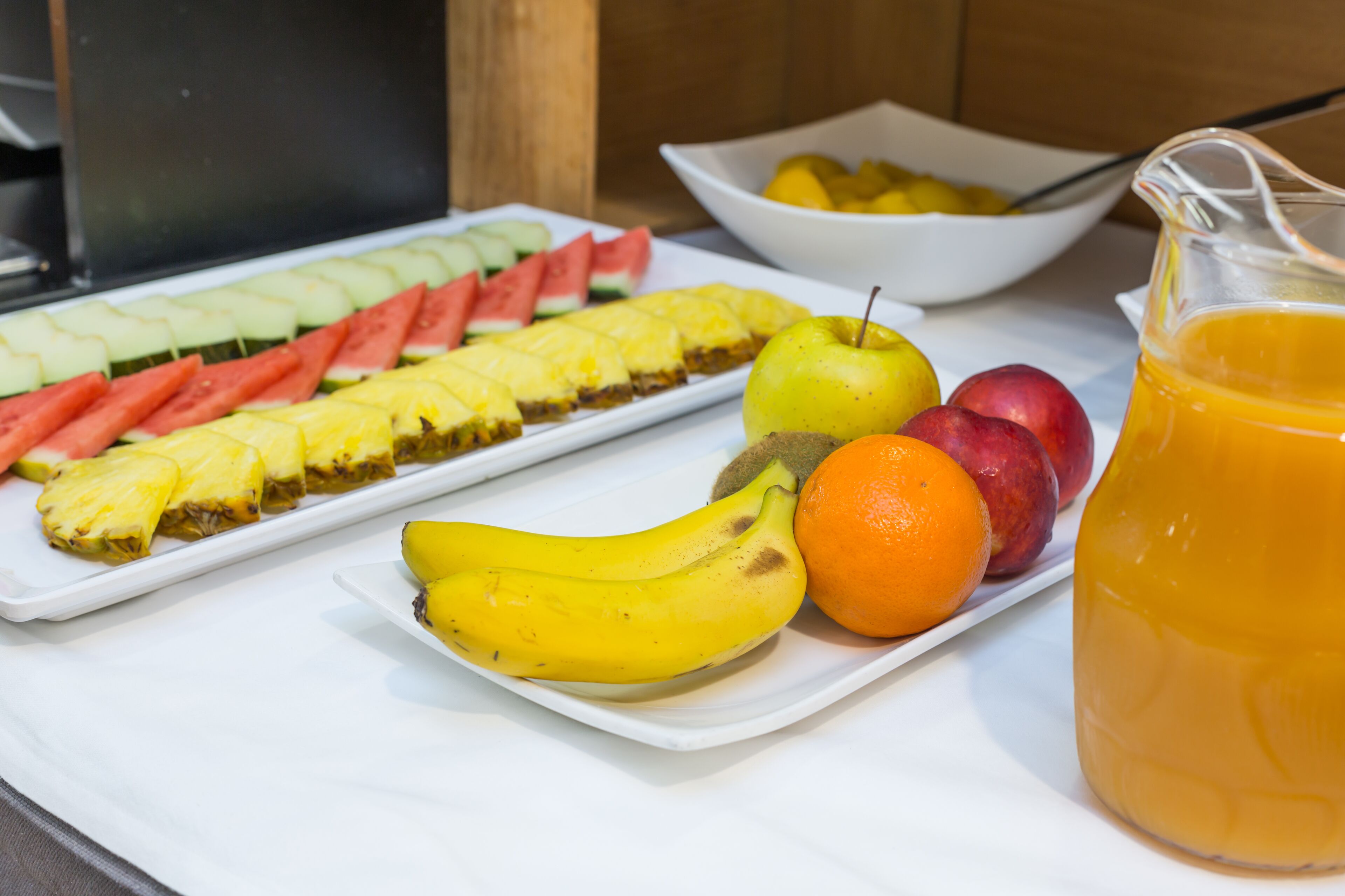 Een ontbijtbuffet met gesneden fruit, een karaf sinaasappelsap en een schaal gemengd fruit.