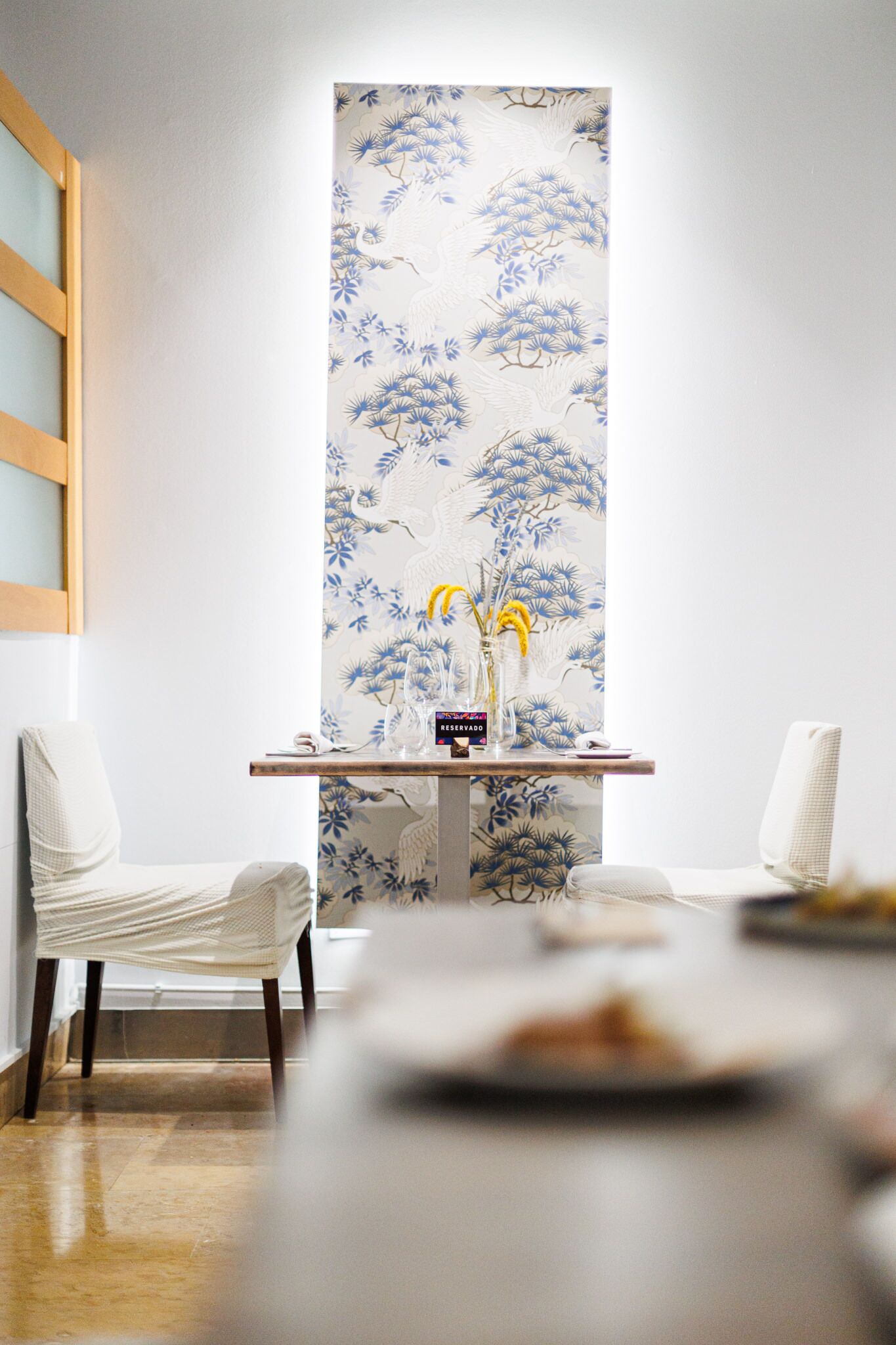 Una habitación con un papel tapiz de estilo tradicional japonés, una pequeña mesa de madera con un jarrón de flores y dos sillas blancas.