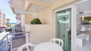 A balcony with a white table and chairs, a glass door leading to a kitchen area, and a view of the street and buildings.