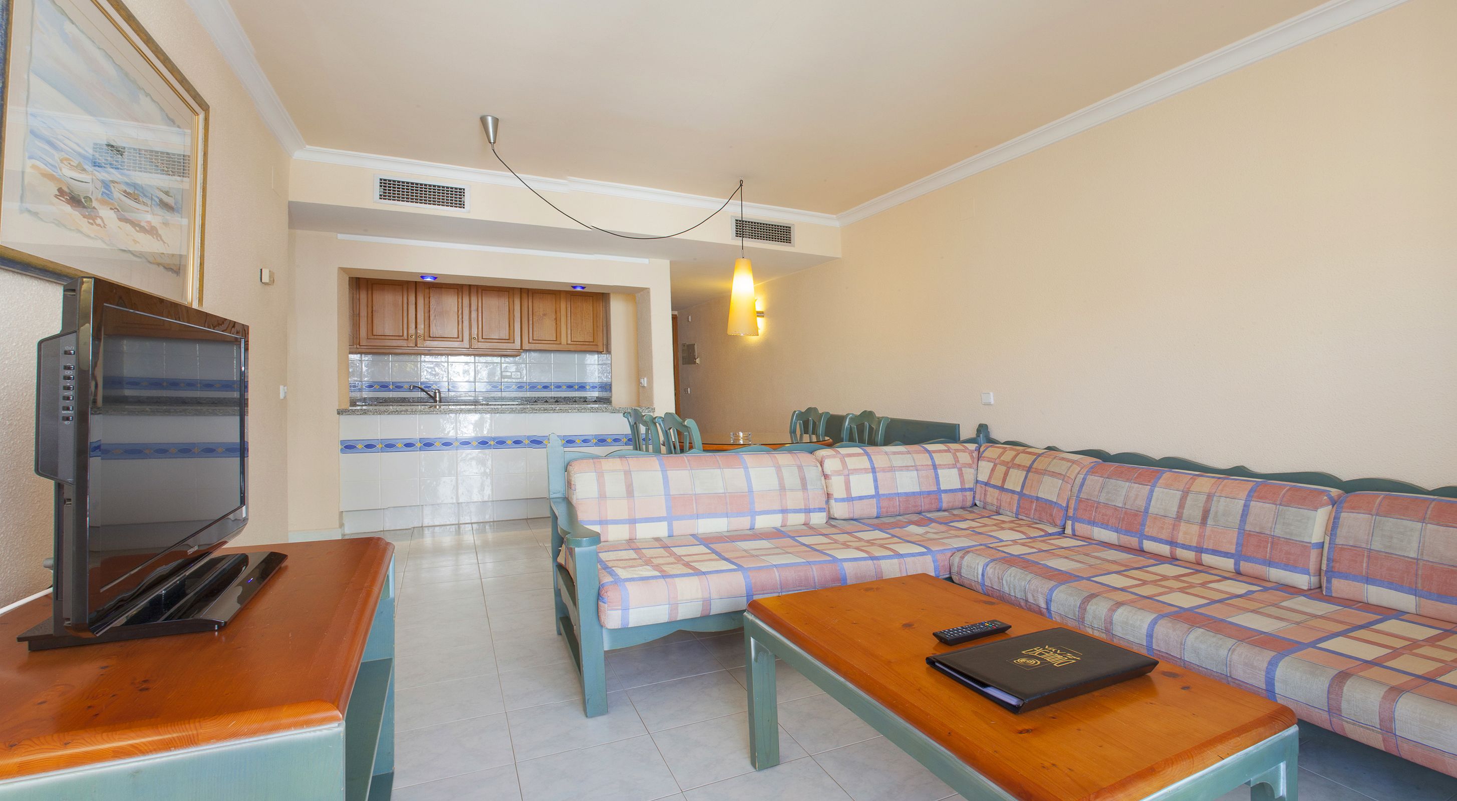 Una sala de estar con un sofá, una mesa de centro y un televisor. Se ve una cocina al fondo con armarios y un lavamanos.