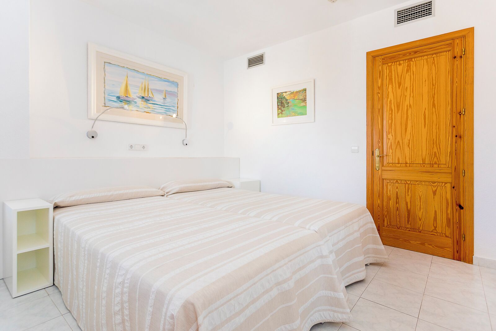 Un dormitorio con cama, mesita de noche, puerta de madera y dos cuadros en la pared.